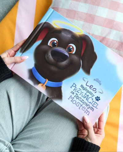 OSOBA TRZYMA KSIĄŻKĘ „LEO, NAJLEPSZY PRZYJACIEL ZA TĘCZOWYM MOSTEM” Z ILUSTRACJĄ PSA Z AUREOLĄ.