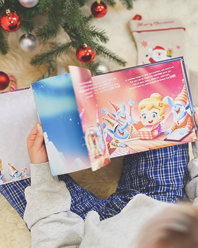 Otwarte spersonalizowane książki świąteczne na kolanach dziecka obok choinki z bombkami i skarpetą na prezenty.